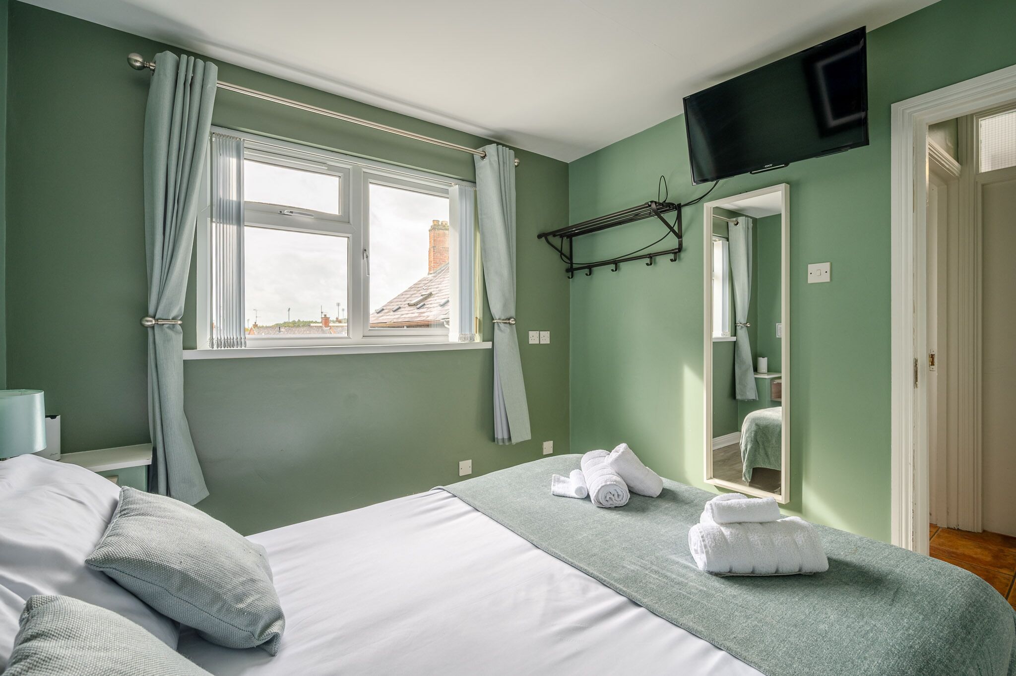 A hotel room with a bed, a TV mounted on the wall, a window with curtains, and a mirror reflecting the room.