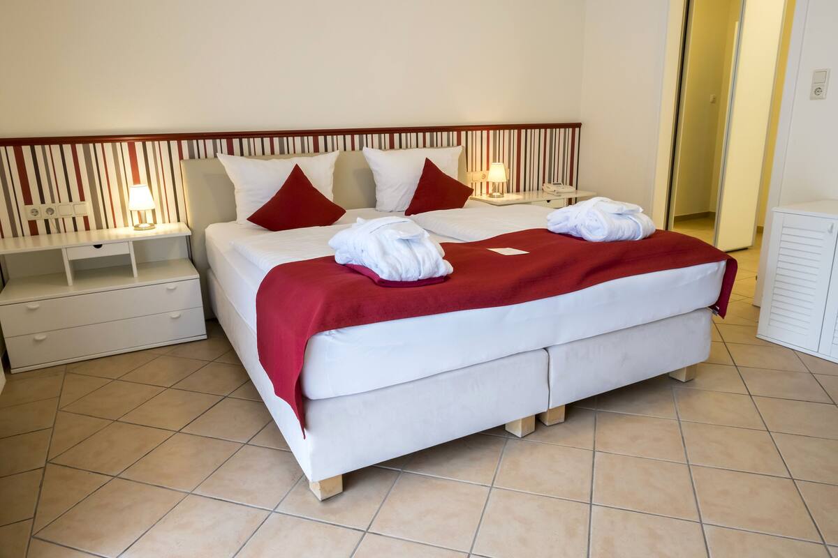 Een net opgemaakt bed met witte lakens en rode kussens, een rode plaid, en twee witte handdoeken op het bed. Er staat een nachtkastje met een lamp bij en een lade onder het bed.