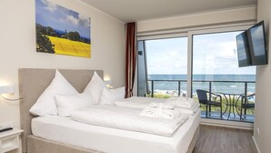 A hotel room with a large bed, a TV mounted on the wall, and a balcony with a view of the sea.