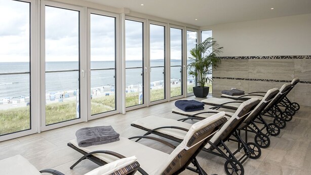 A sunlit room with large windows overlooking a beach, featuring lounge chairs and a potted plant.