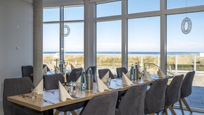 A dining area with a long table set for six, large windows offering a view of the beach and dunes, and a modern interior design.