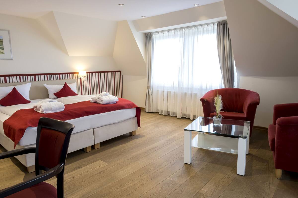Een moderne hotelkamer met een bed, rode stoelen, een glazen bijzettafeltje en een groot raam met gordijnen.