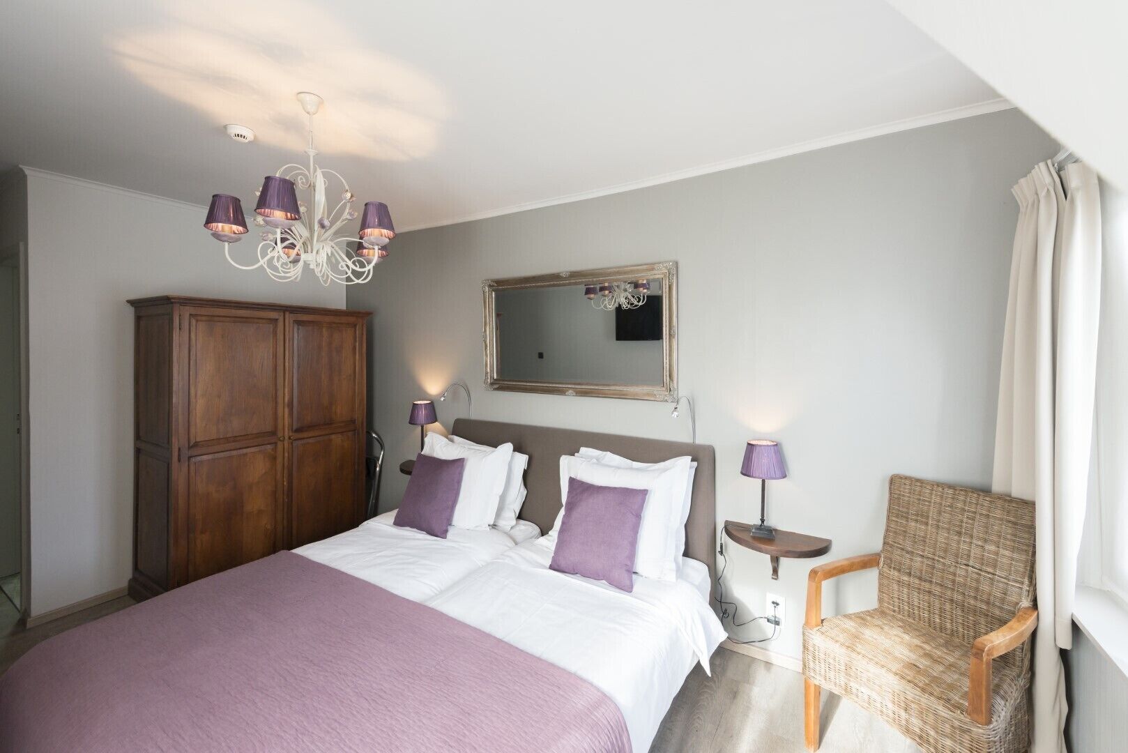 Een slaapkamer met een groot bed, een houten garderobe, een rieten stoel, een spiegel en een kroonluchter.