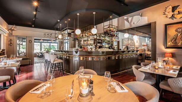 A restaurant with a bar area, round tables with candles, and a view of the outdoors.
