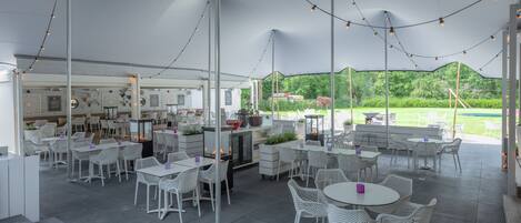 Un espace de restauration en plein air avec des tables et des chaises en osier blanc, des guirlandes lumineuses suspendues et une vue sur la verdure en arrière-plan.