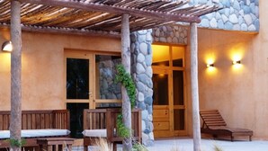 Katettu ulkoalue, jossa on puiset penkit ja pergola, kiviseinä sekä lasipaneeleilla varustettu puuovi.