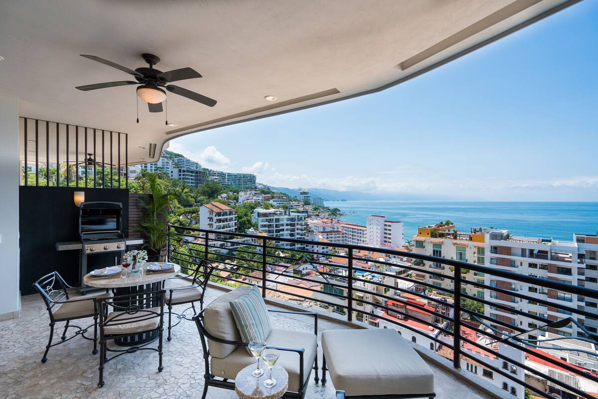 A balcony with a grill, seating, and a view of the city and ocean.