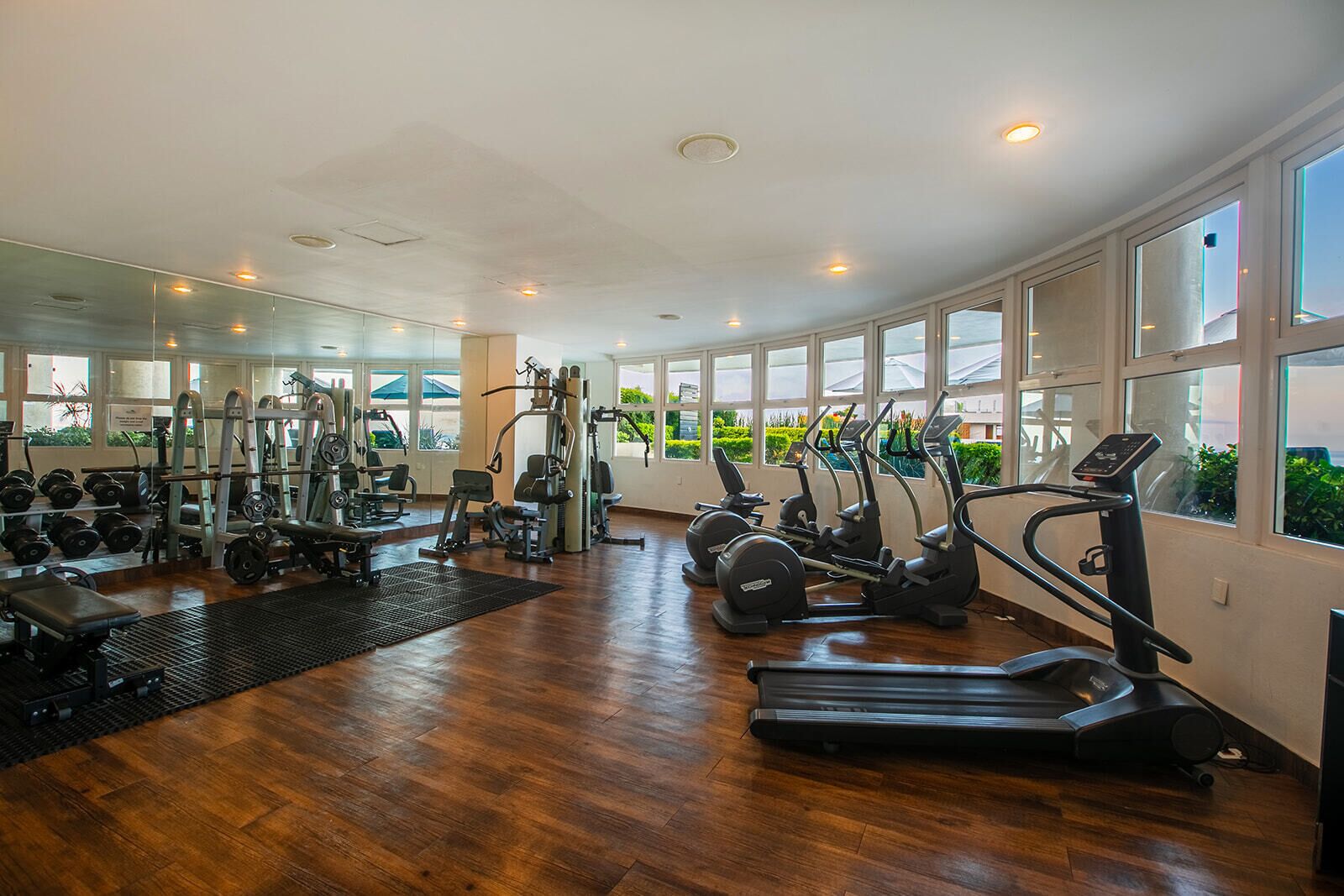 A spacious gym with various exercise equipment, large windows, and a wooden floor.