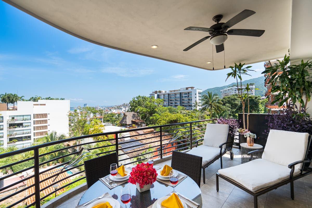A balcony with a table set for two, a ceiling fan, and a view of the cityscape.
