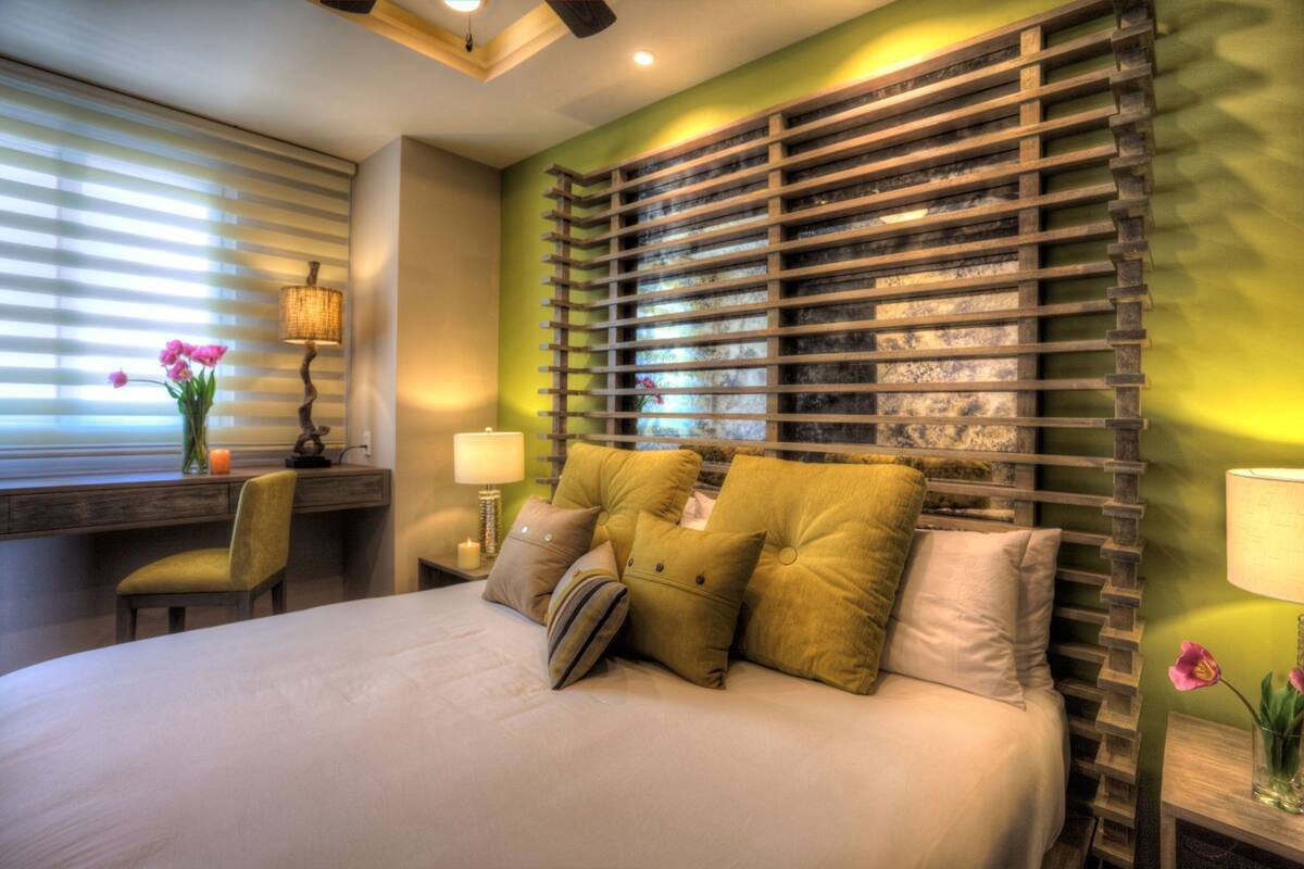 A bedroom with a bed, a chair, a nightstand, a lamp, and a window with blinds.