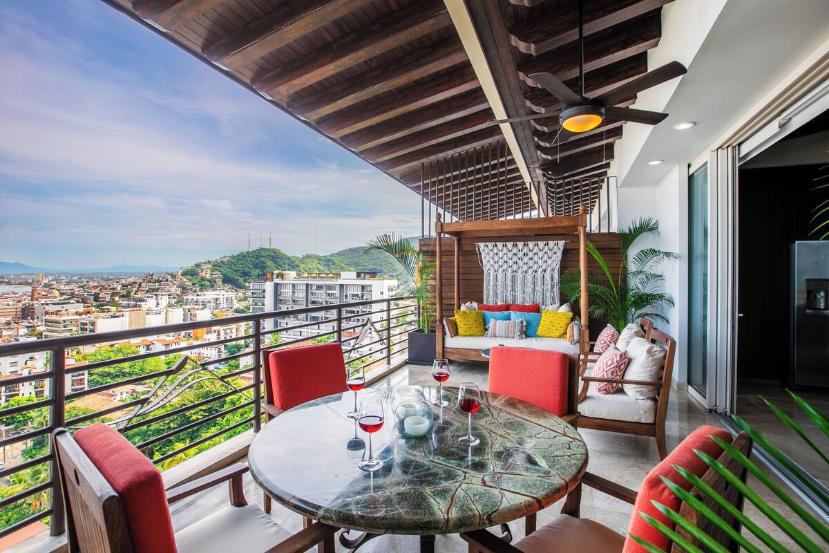 A balcony with a glass table, red chairs, and a sofa with colorful cushions.