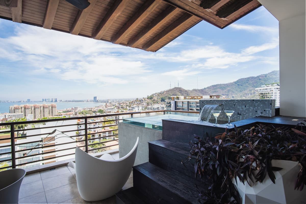 A balcony with a view of the city, a pool, and modern furniture.