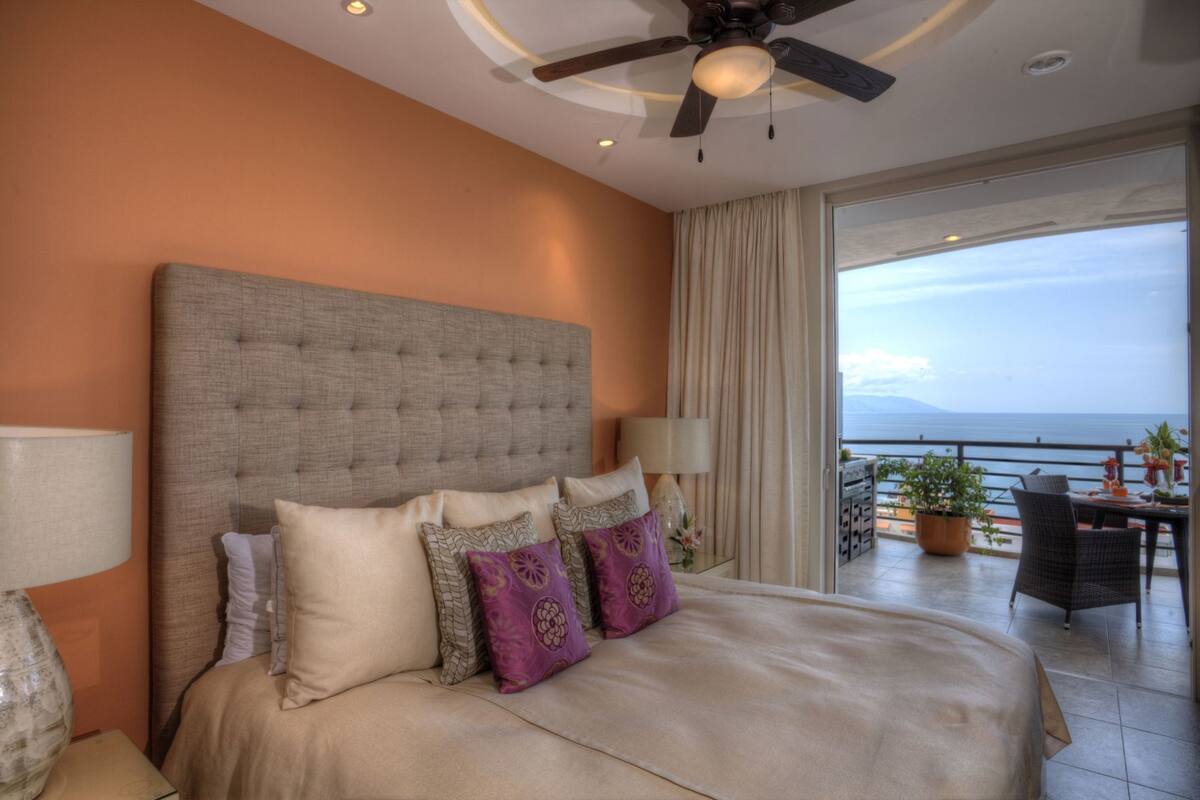 A bedroom with a large bed, a ceiling fan, a view of the sea, and a balcony with outdoor furniture.
