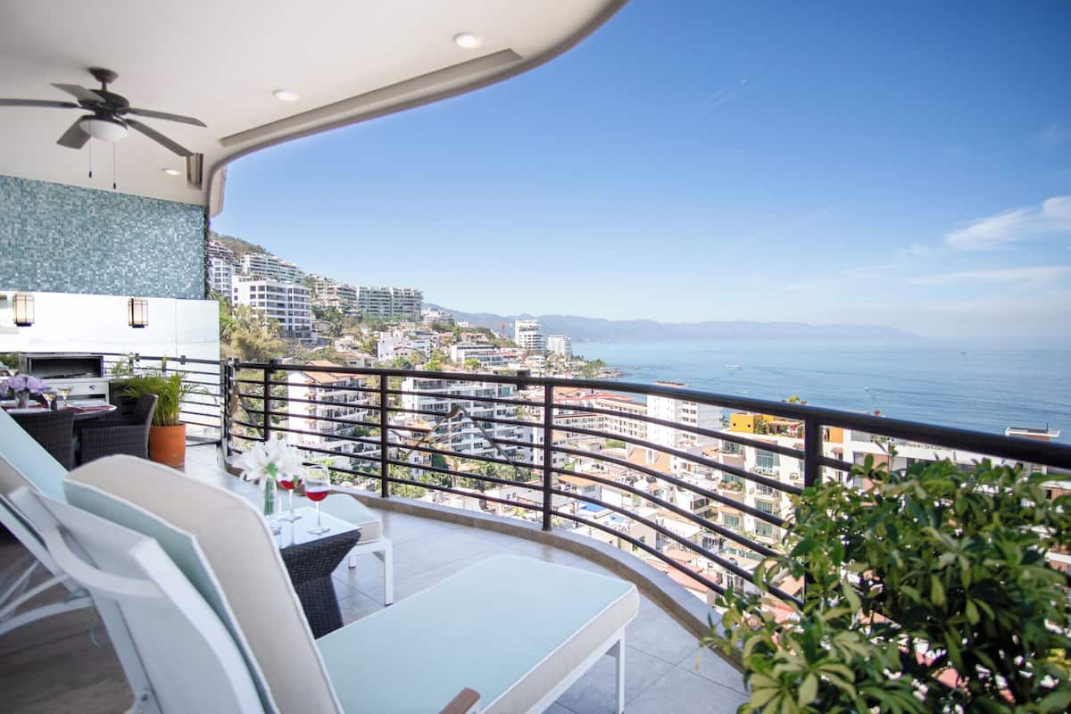 A balcony with a view of the sea and cityscape, furnished with chairs and a table, and a ceiling fan.