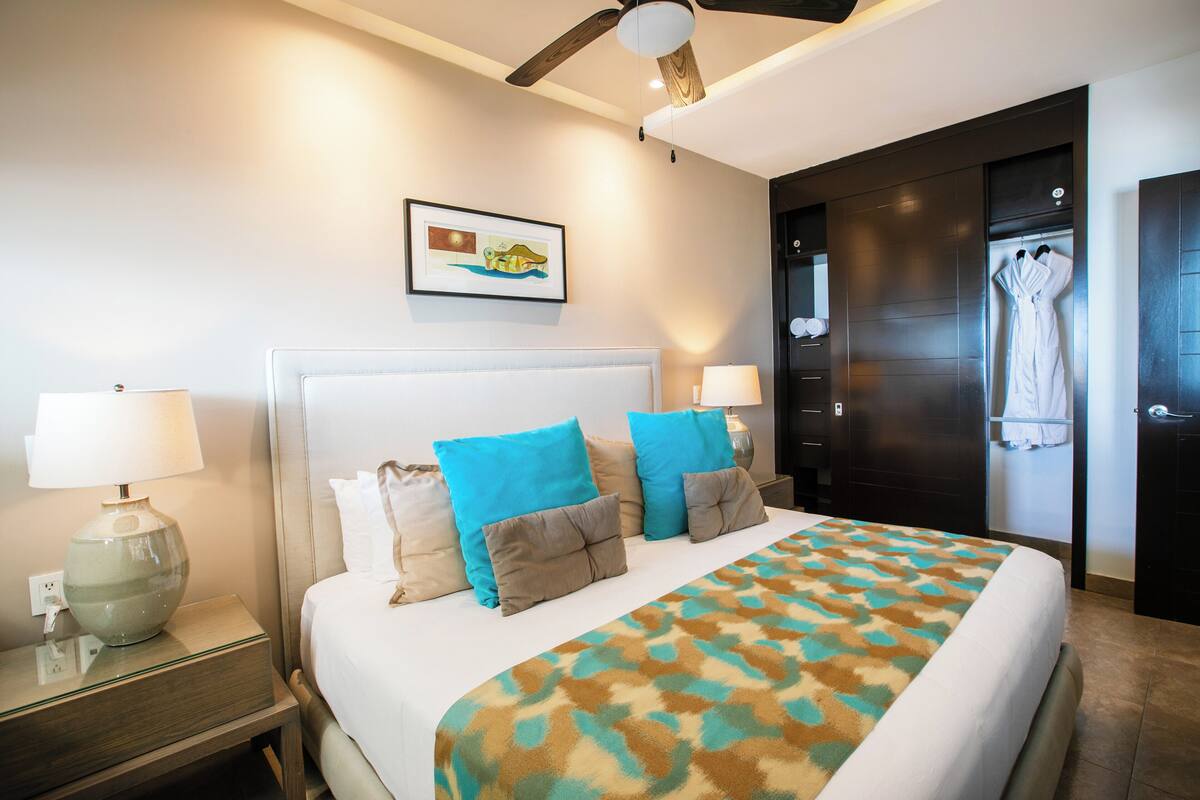 A hotel room with a large bed, two bedside lamps, a ceiling fan, a framed picture on the wall, and a door with a towel rack.