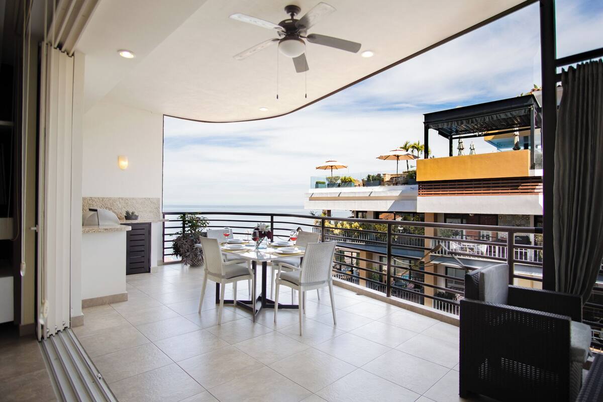 A balcony with a dining table, chairs, and umbrellas, overlooking a coastal view.