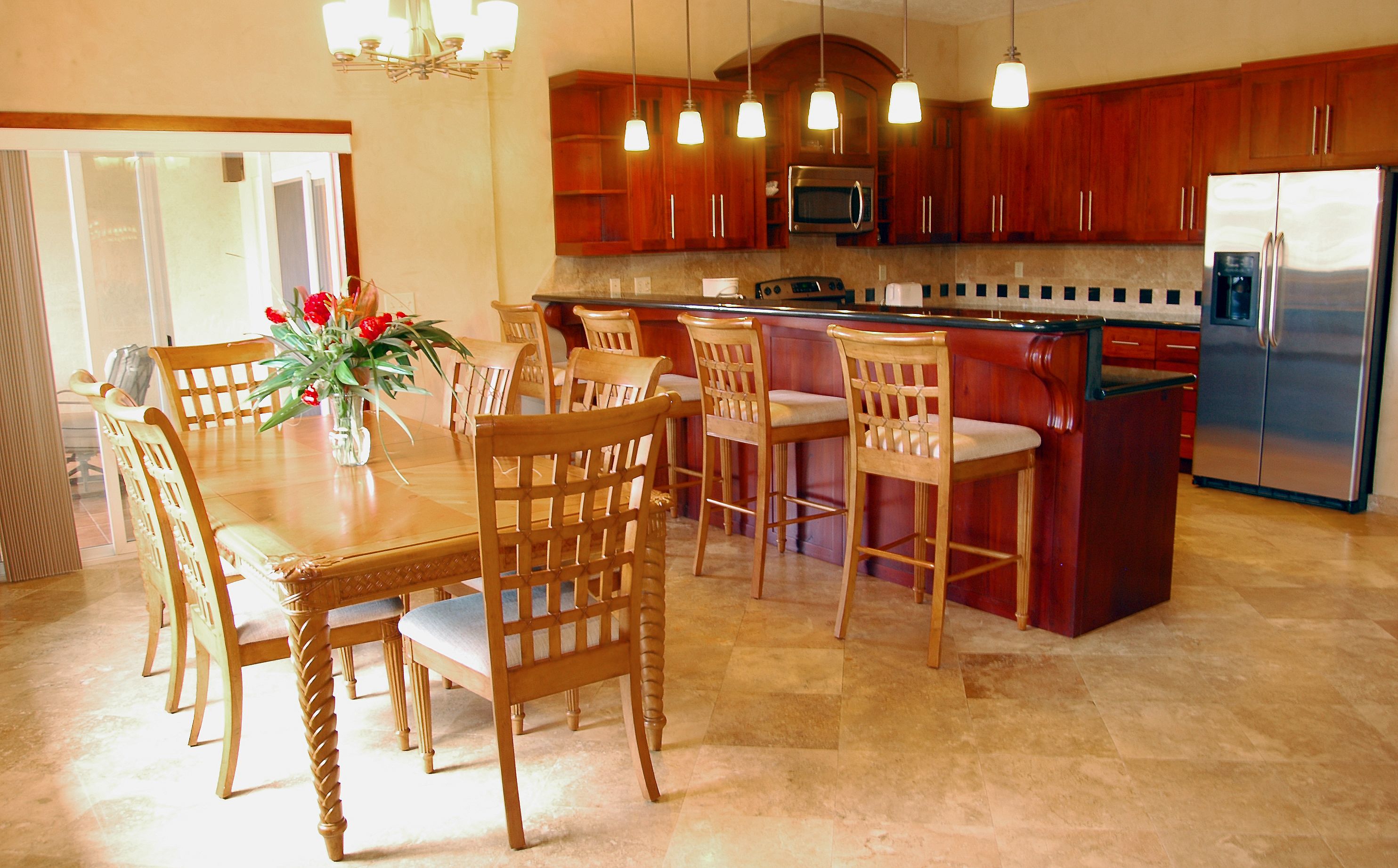 A kitchen with wooden cabinets, a stainless steel refrigerator, and a dining area with wooden chairs and a vase of flowers.