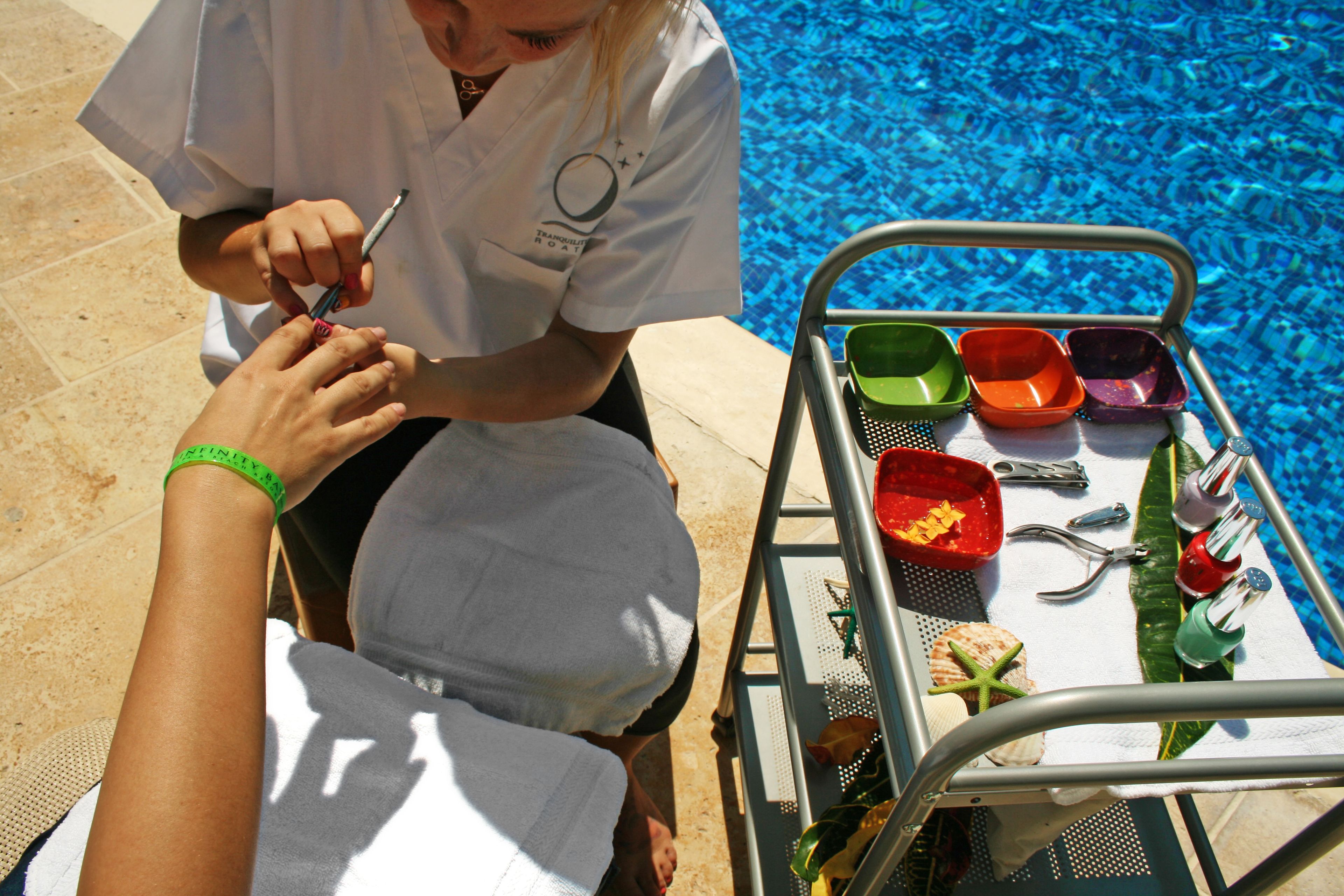 A person receiving a manicure by a professional near a poolside area with a metal cart containing various items.