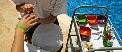 A person receiving a manicure by a professional near a poolside area with a metal cart containing various items.