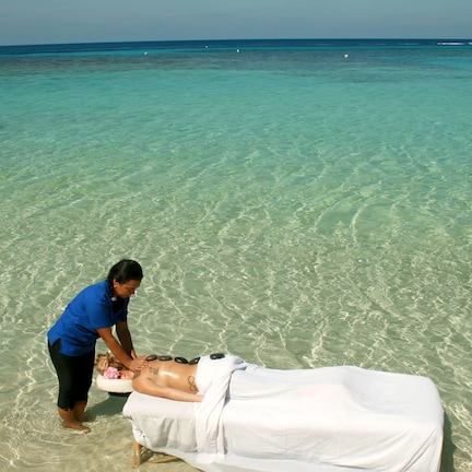 A person receiving a massage on a beach with clear turquoise water and a boat in the distance.