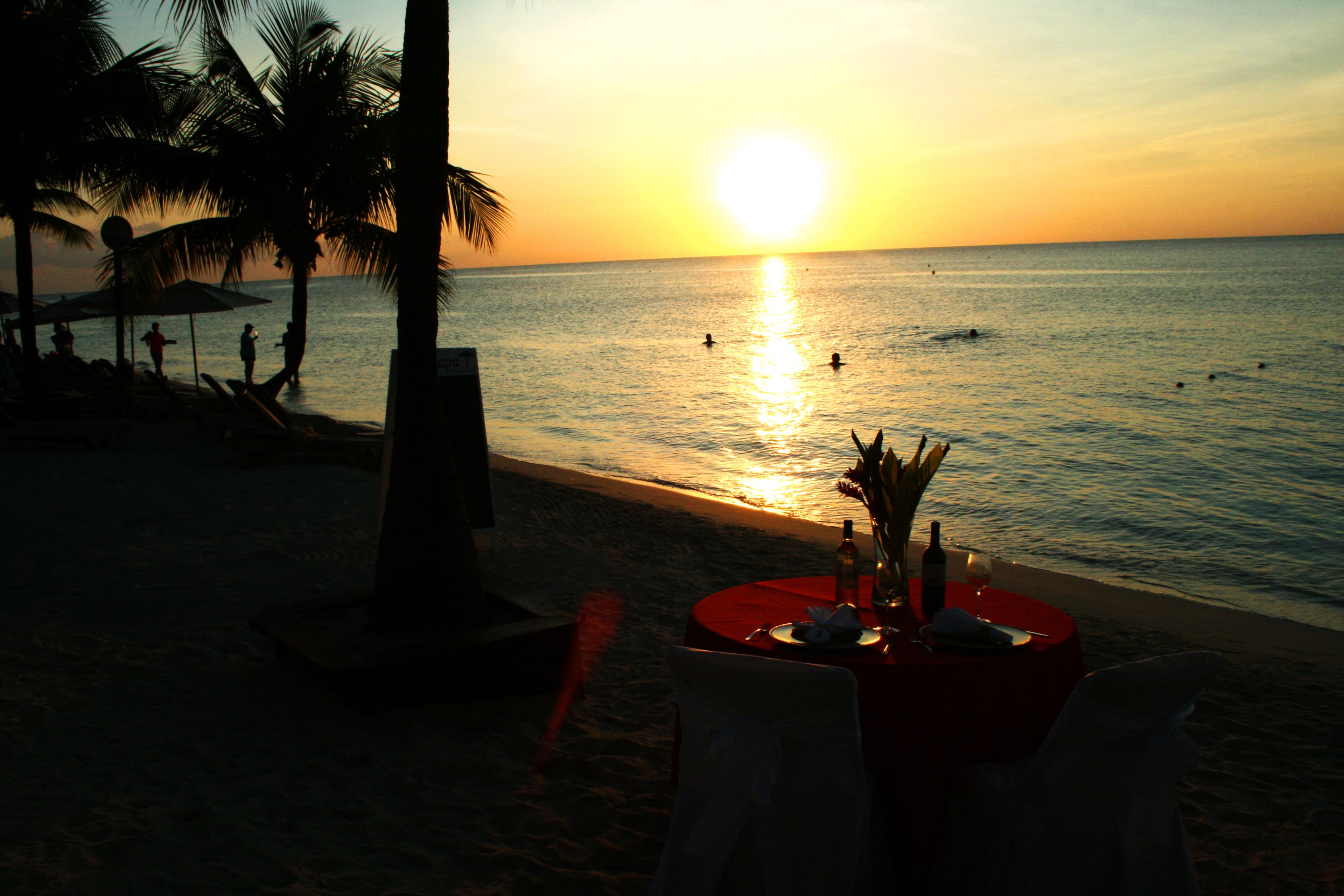 Mesa en la playa con mantel rojo, una botella y un vaso, sobre un fondo de atardecer.