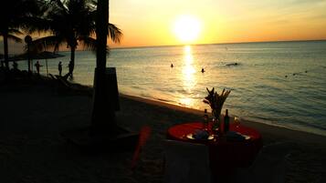 Mesa en la playa con mantel rojo, una botella y un vaso, sobre un fondo de atardecer.