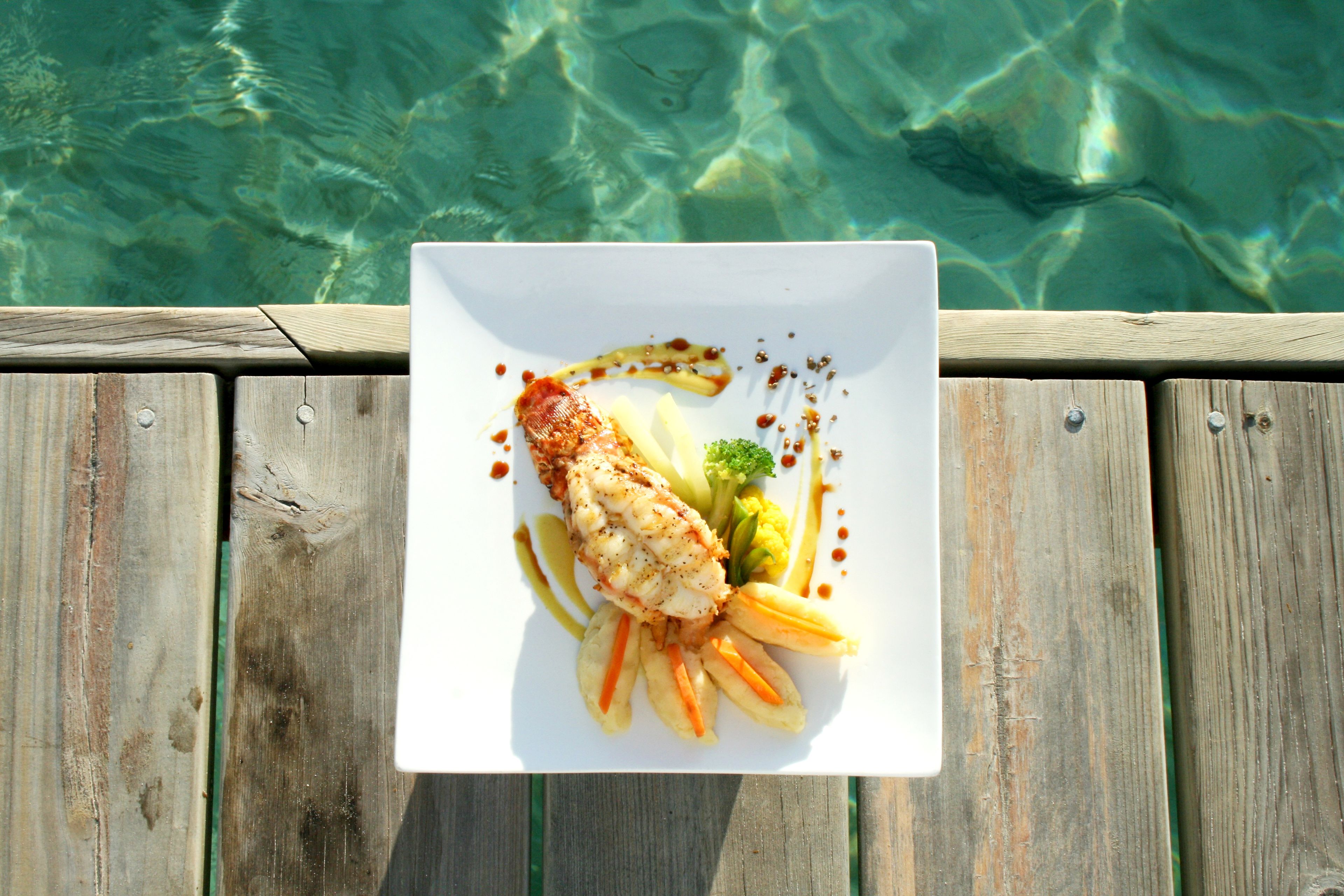Une assiette blanche contenant du poisson, des légumes et de la sauce, posée sur une terrasse en bois, au bord d’une étendue d’eau claire.