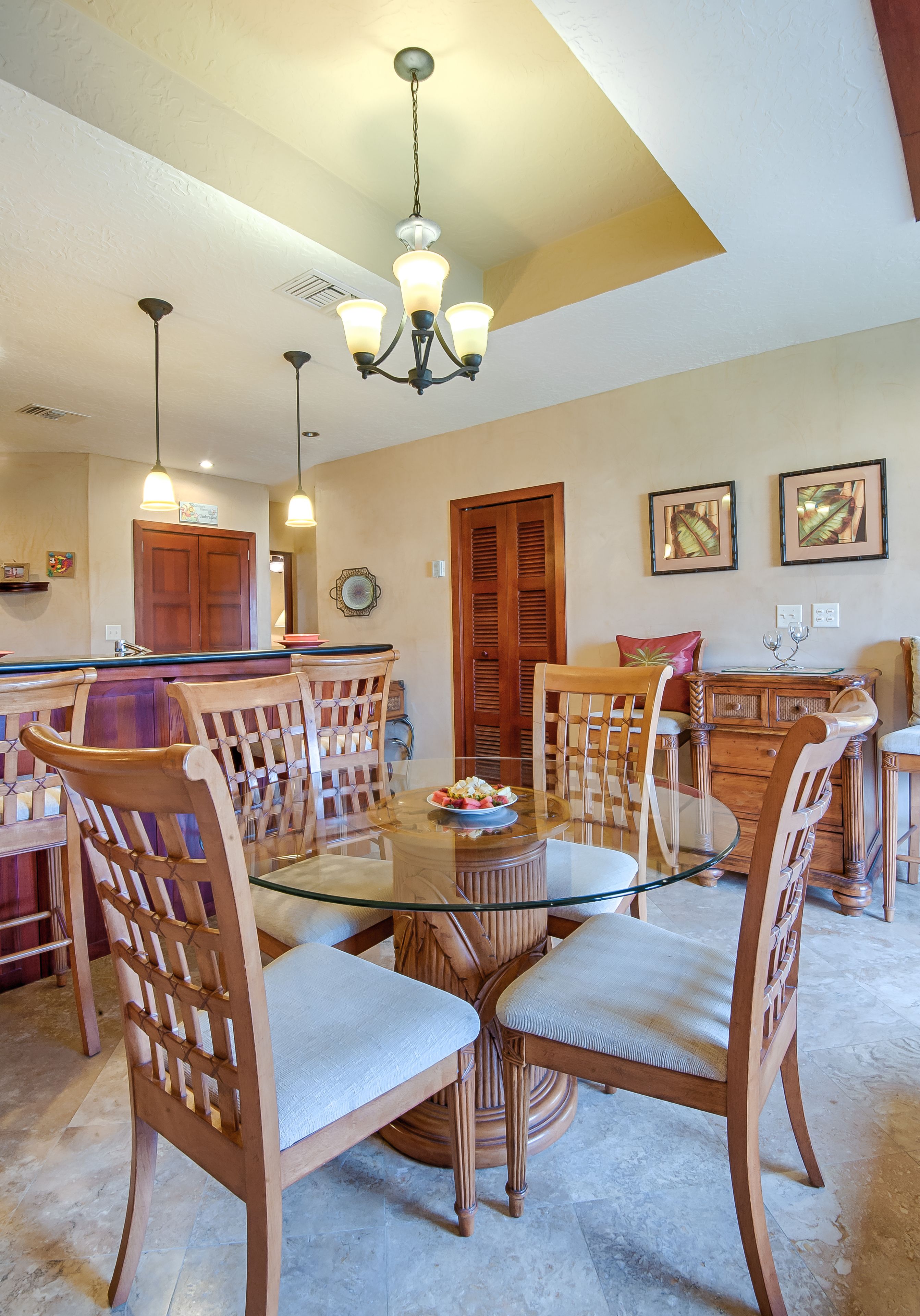 A dining area with a glass-top table surrounded by wooden chairs, a chandelier, and wooden cabinets.