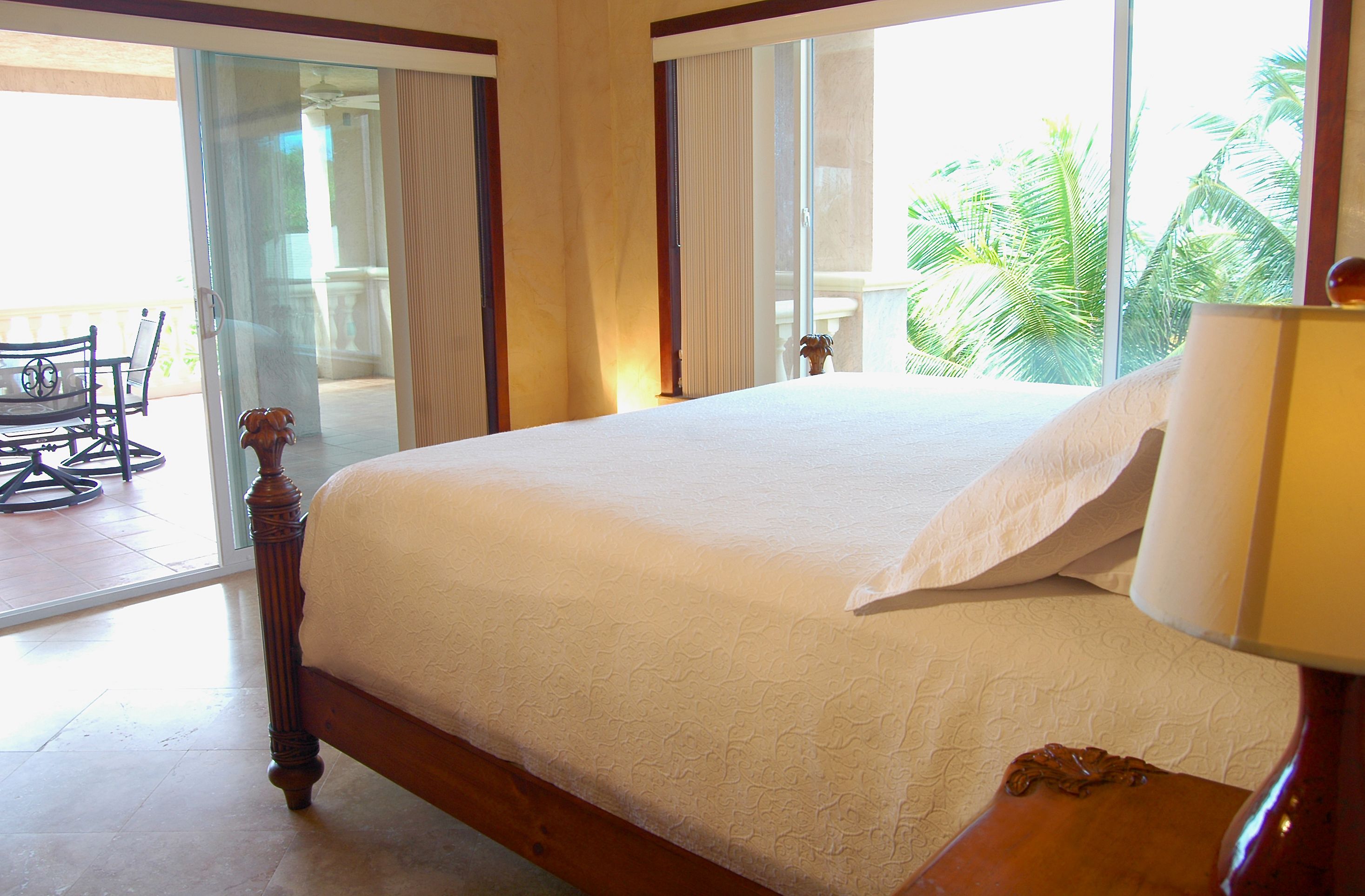 Uma cama grande com estrutura de madeira e roupa de cama branca, uma mesinha de cabeceira de madeira com um abajur, e vista para uma varanda com cadeira e palmeiras.