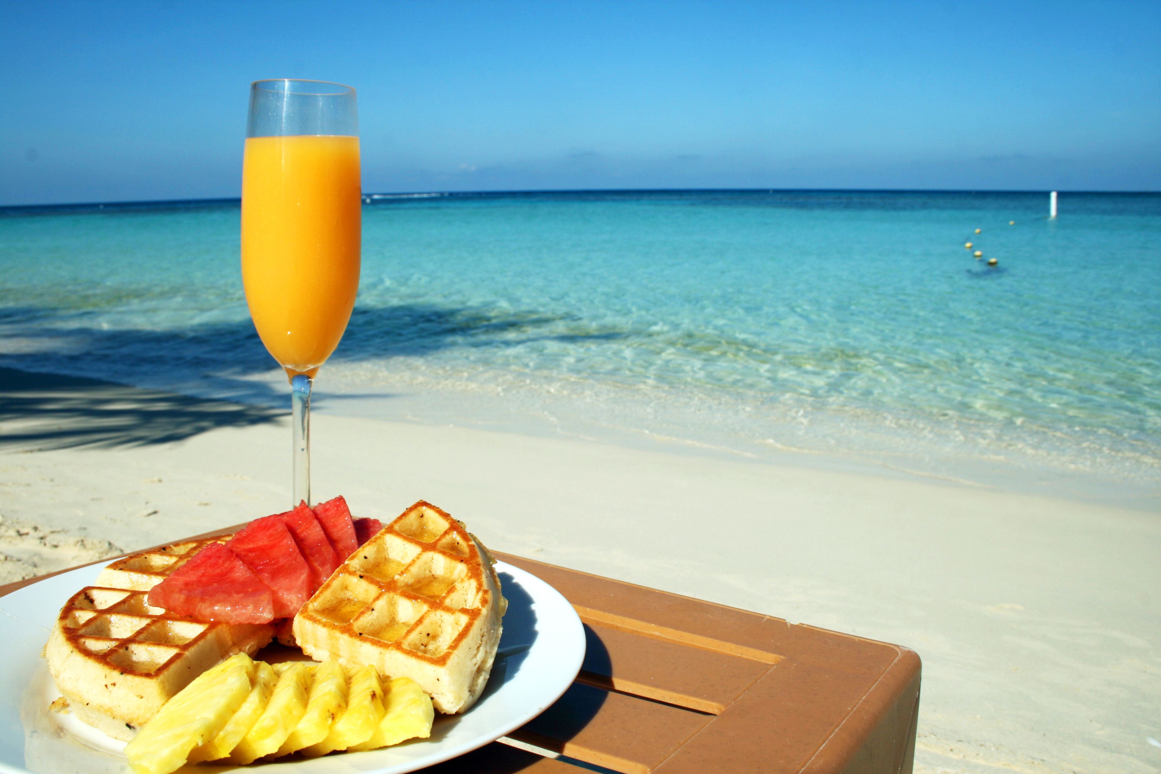 A plate with waffles, watermelon, and pineapple on a beach with clear water and a clear sky.