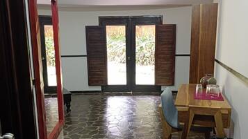 Ein Zimmer mit Steinboden, einem Holztisch mit Stühlen, einer Tür mit roten Vorhängen und einem Ausblick durch ein Fenster mit Holzläden.