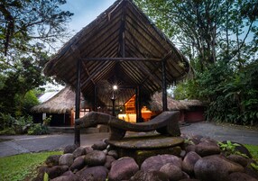 Uma construção com telhado de palha e uma escultura de pedra em primeiro plano, rodeada por rochas e vegetação.