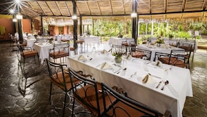 Un restaurant doté de nombreuses tables dressées pour le service, d’un toit de chaume et offrant une vue sur la verdure environnante.