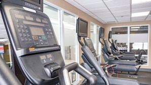 Une rangée de vélos d’appartement fixes, dotés d’écrans numériques et de panneaux de commande, est disposée dans une salle de sport avec de grandes fenêtres et un plafond carrelé.