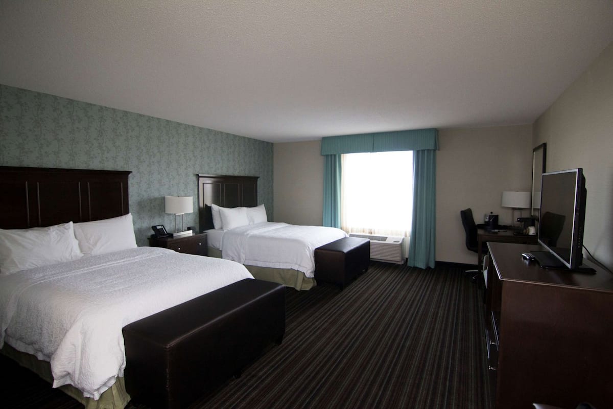 A hotel room with two beds, a desk with a TV, a chair, and a window with curtains.