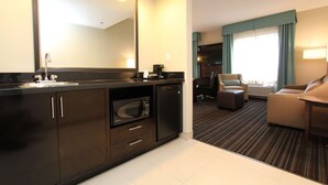 Une chambre d’hôtel dotée d’une kitchenette, d’un grand miroir, d’un coin salon avec un canapé et un fauteuil, et offrant une vue sur l’extérieur par une fenêtre.