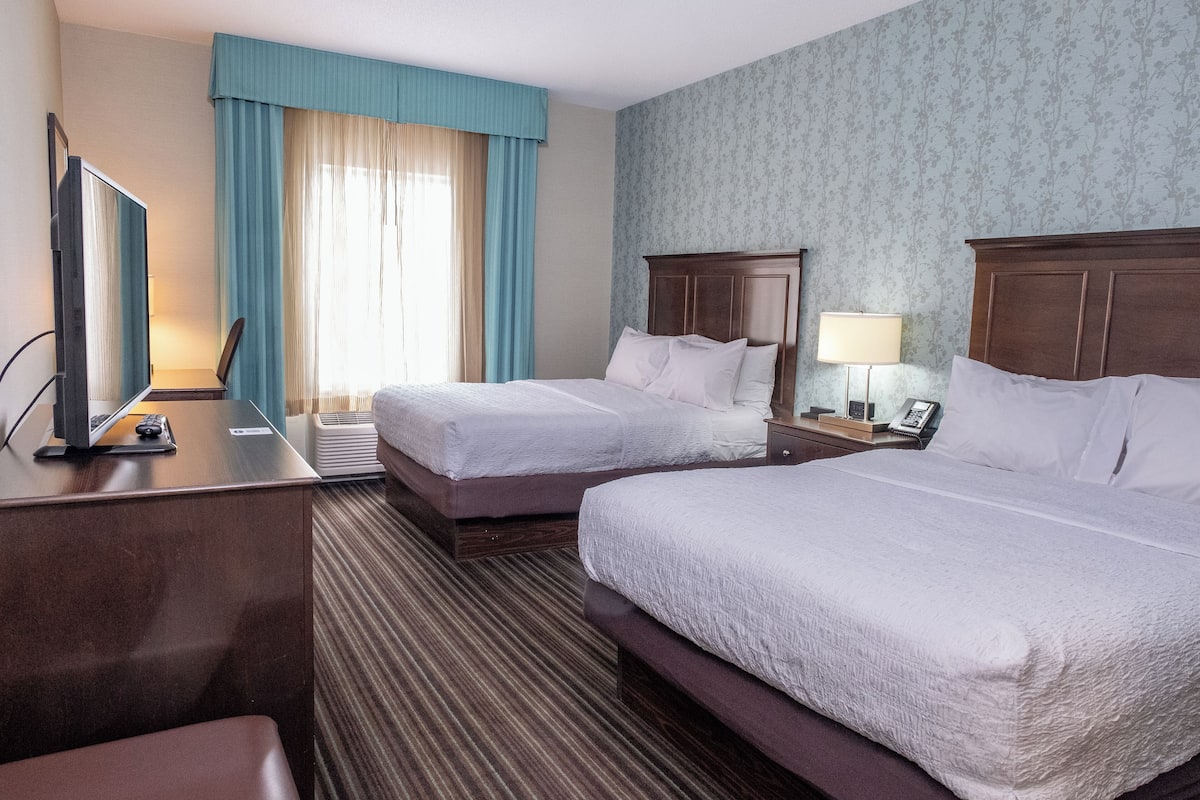 A hotel room with two beds, a desk, a chair, a TV, and a window with curtains.