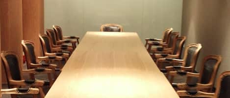 Un salón de conferencias con una larga mesa de madera y sillas, iluminación empotrada en el techo y una pared lisa.