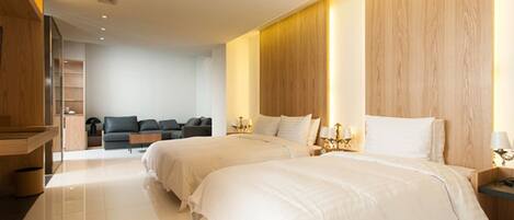 A modern hotel room with two beds, a wooden headboard, a sofa, and a desk with a lamp.