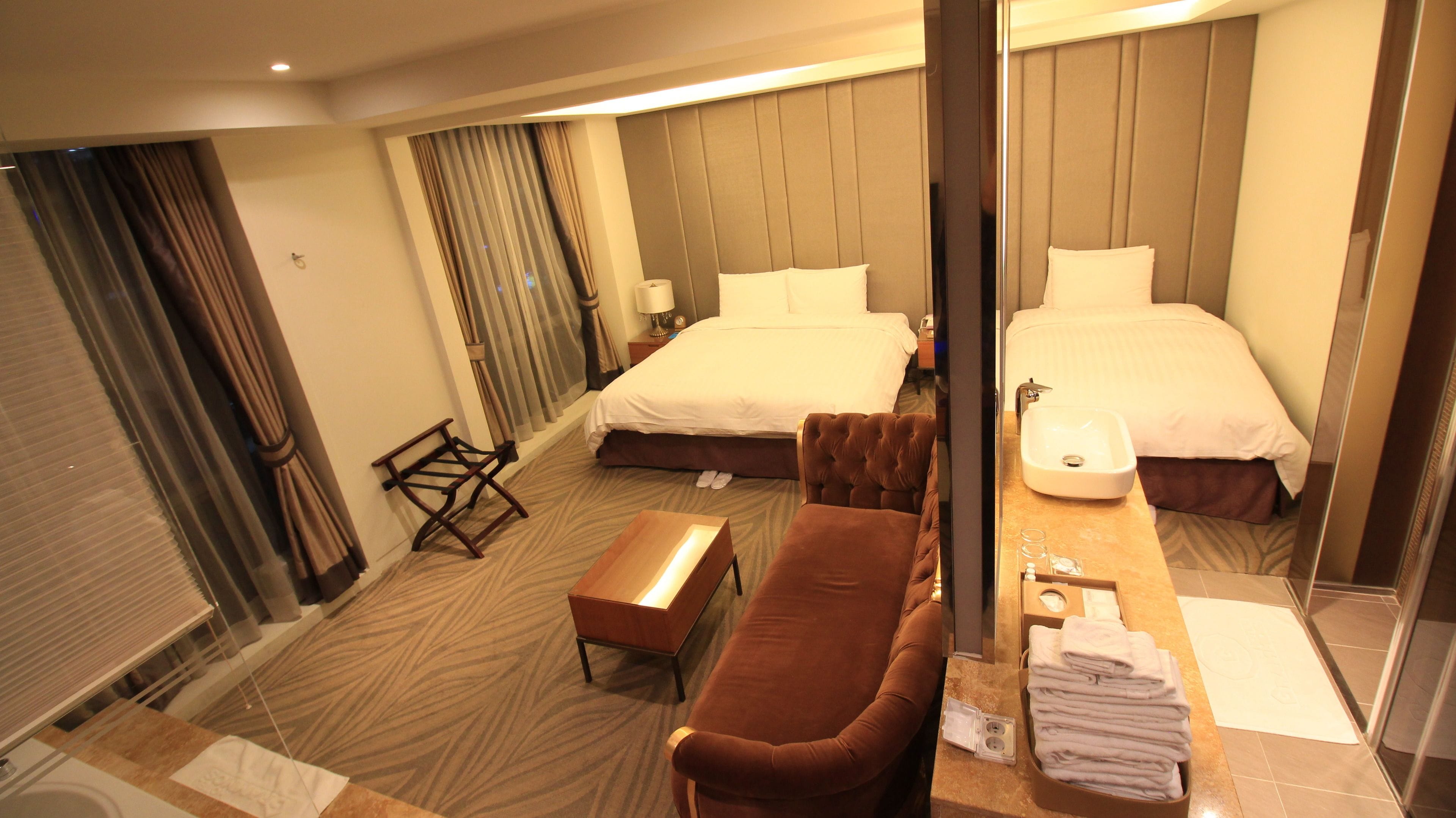 A hotel room with two beds, a sofa, a small table, a mirror, and a bathroom with a sink and towel rack.