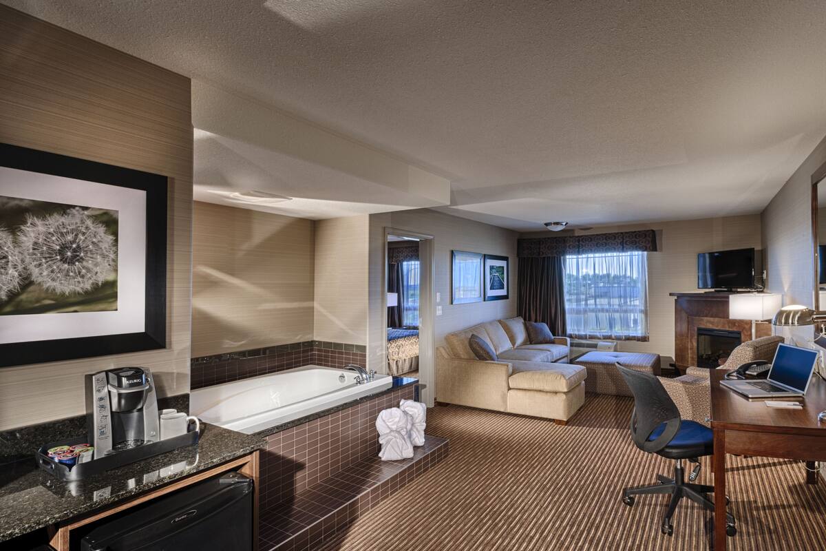 A hotel room with a kitchenette, a large bed, a sofa, a desk with a laptop, a TV, and a fireplace.