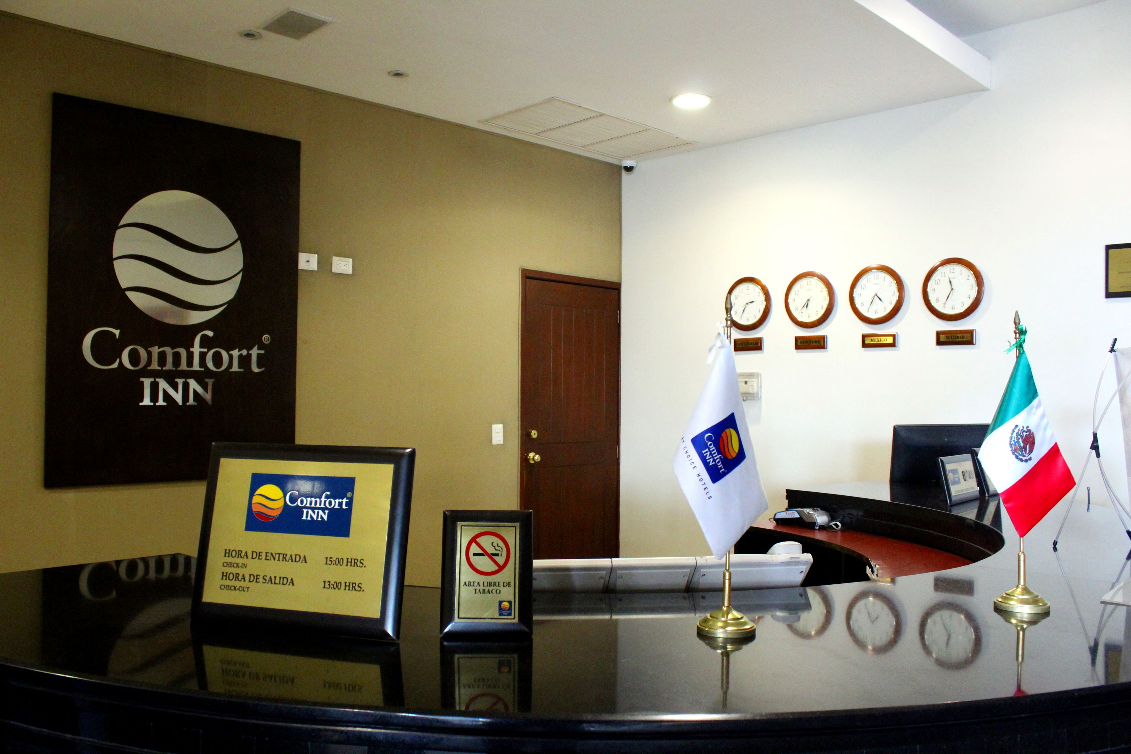 A reception desk at Comfort Inn with a sign indicating check-in and check-out times, a no-smoking sign, and multiple clocks on the wall.