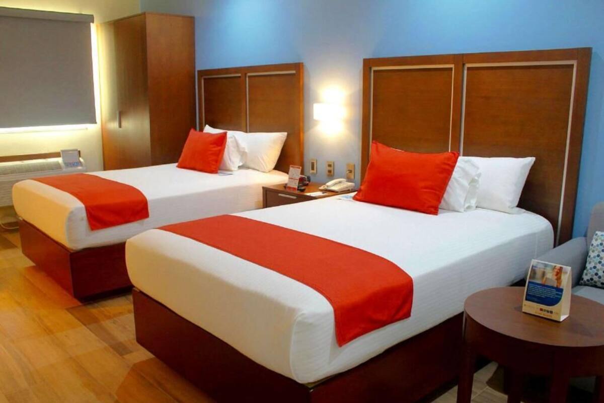 Habitación de hotel con dos camas, cada una con una colcha roja, cabecero de madera, mesita de noche con lámpara y una pequeña mesa redonda con un folleto.