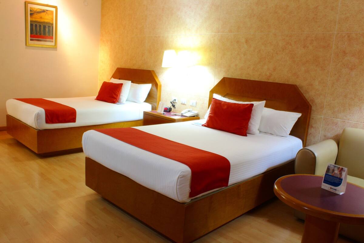 Una habitación de hotel con dos camas, cada una con colcha roja y blanca, una mesita de noche con lámpara y una pequeña mesa con un cartel.
