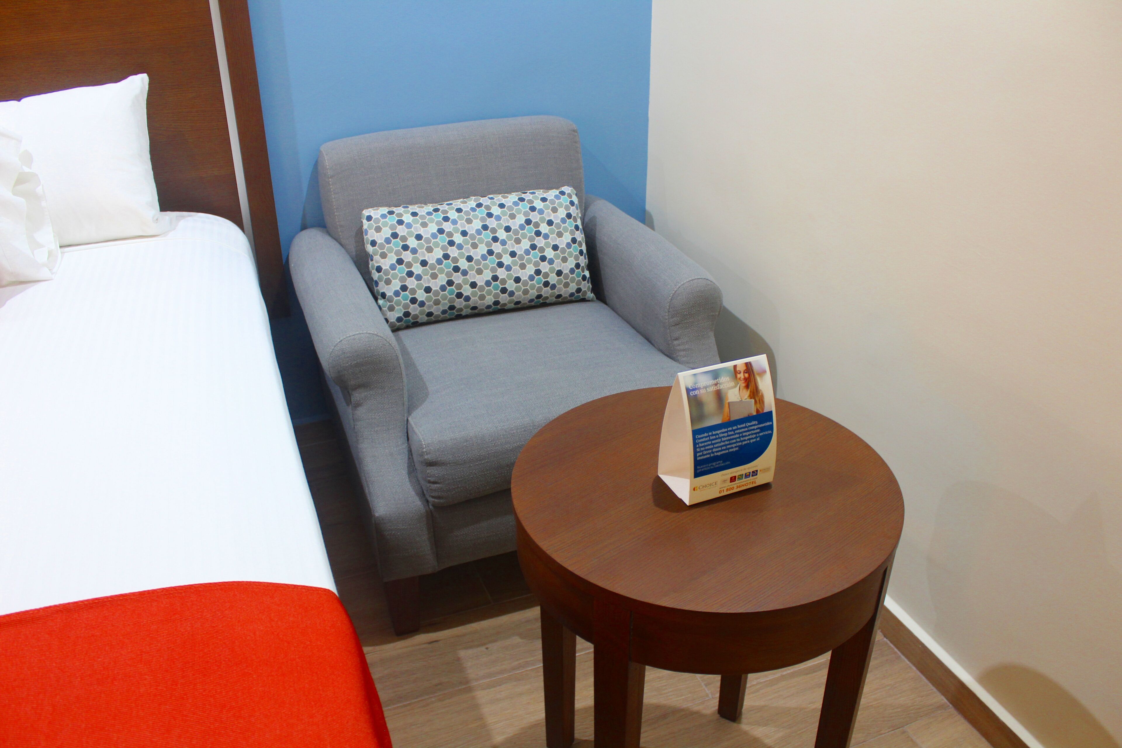 A hotel room with a bed, a grey armchair, a small wooden table, and a pillow with a blue and white pattern.