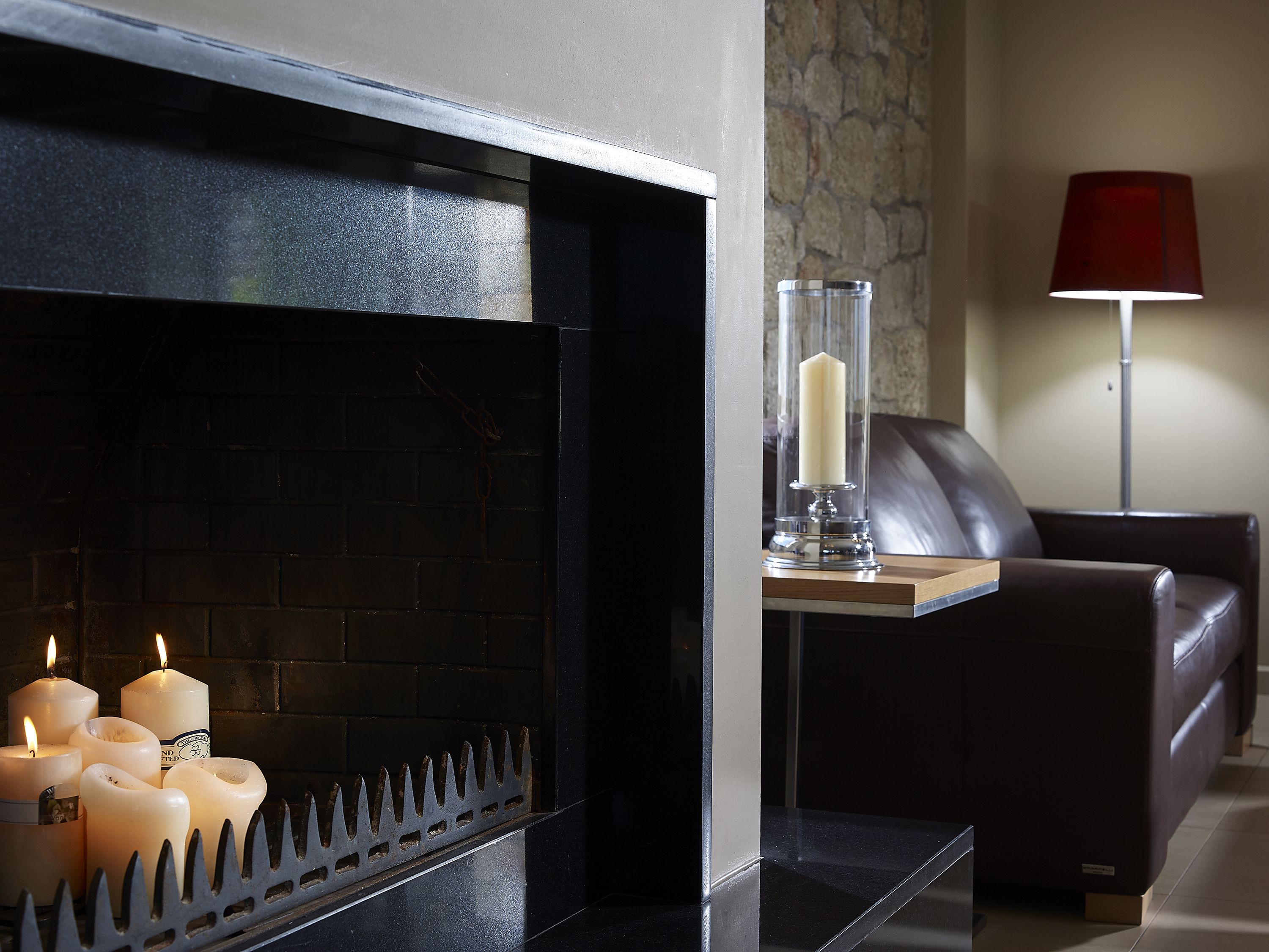 A fireplace with candles, a glass candle holder, and a red lamp on a table.
