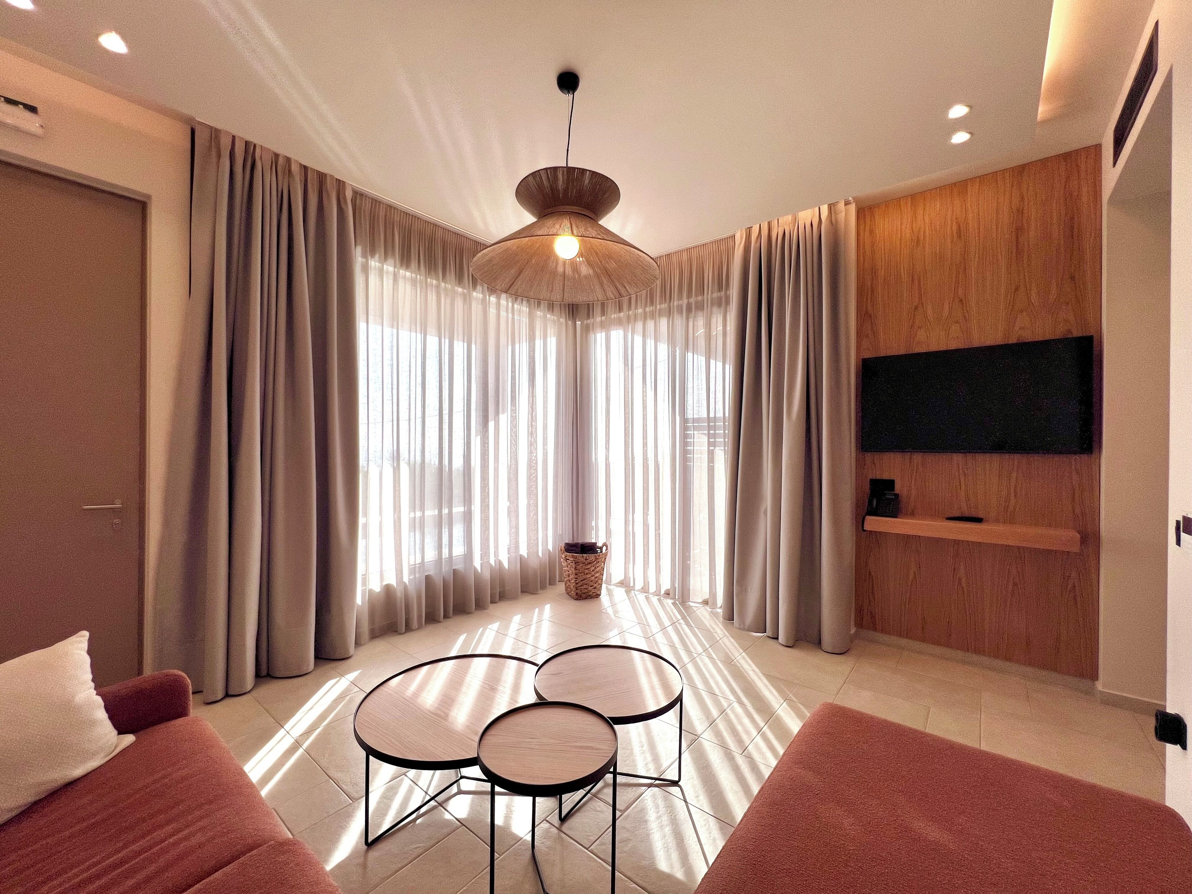 A modern living room with a sofa, two round tables, a TV mounted on the wall, and a large window with curtains.