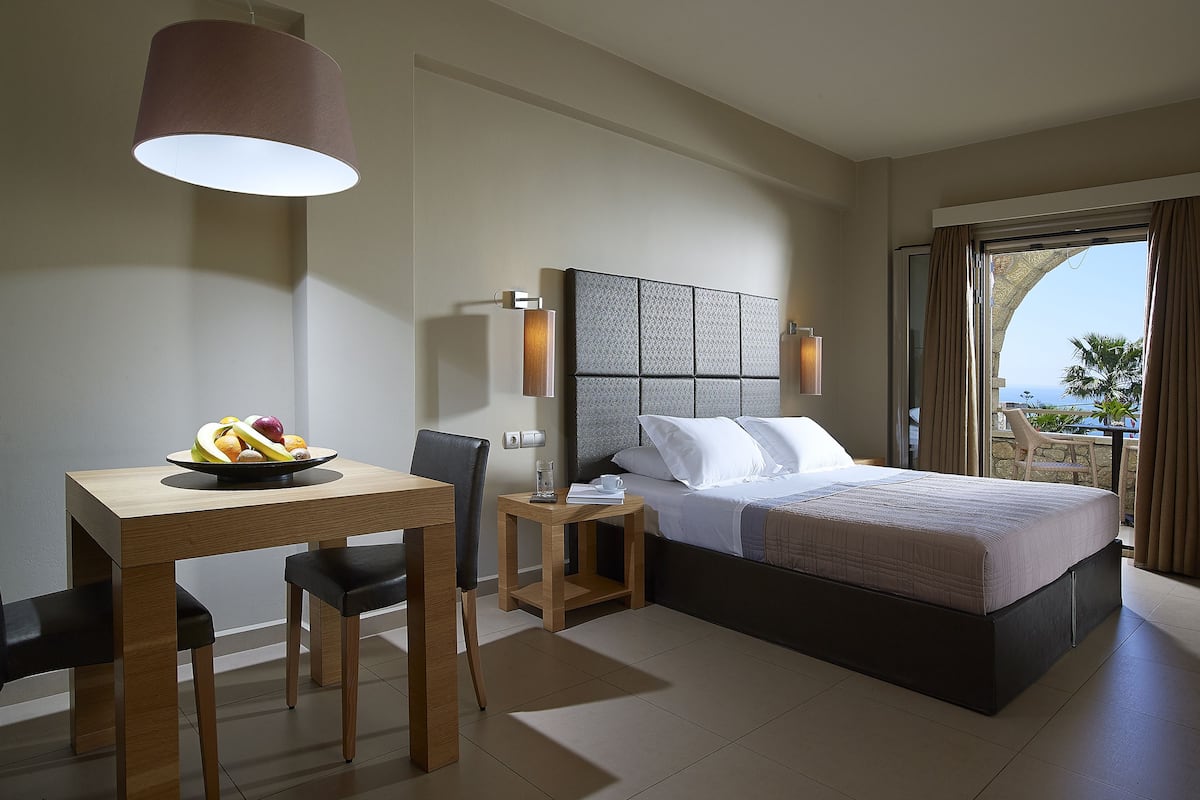 Ein modernes Hotelzimmer mit einem großen Bett, einem Esstisch mit Stühlen, einem kleinen Beistelltisch und Blick aufs Meer.