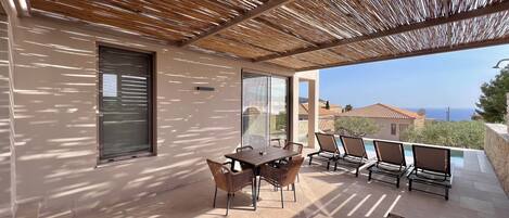 A covered outdoor area with a table and chairs, a poolside seating area, and a view of the sea.