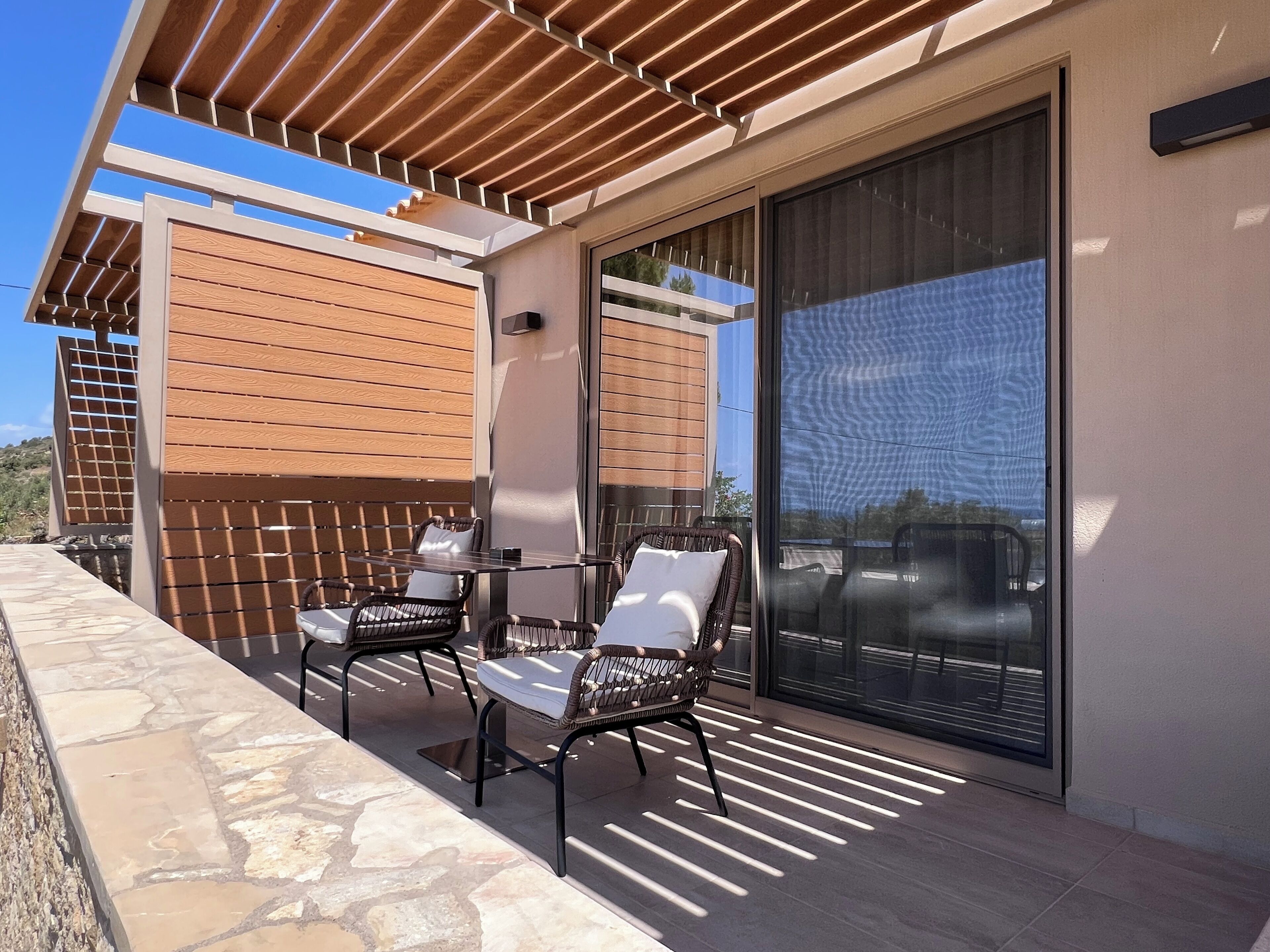 A balcony with two wicker chairs and a table, a pergola overhead, and a large sliding glass door.